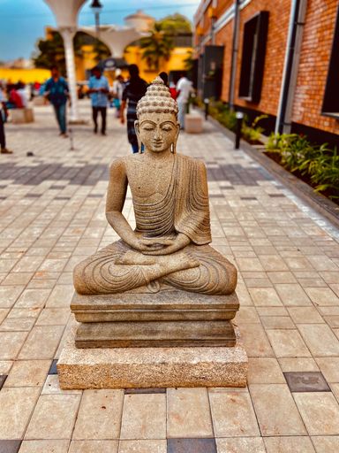 A serene stone statue of the Buddha is situated on a raised platform in a bustling cityscape