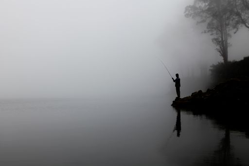 A silhouette of a person fishing near a lake with reflection and surrounded by fog and mist