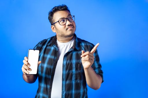 portrait of an Indian man showing smart phone with white screen and pointing finger aside on blue background