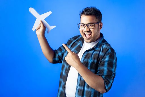 portrait of a happy young man holding paper cut airplane and pointing with finger, smiley face, isolated on blue background