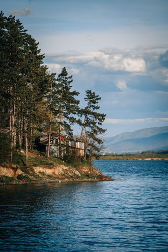 A vertical stock photo of a quaint house located near a tranquil lake, surrounded by lush greenery and a beautiful blue sky