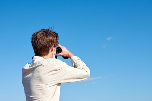 A close up of a young man in a white shirt looking through binoculars on blue space -view from the back