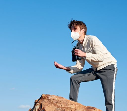 A low angle view of a young man from Spain with binoculars wearing a mask standing on a mountaintop astonished by the view