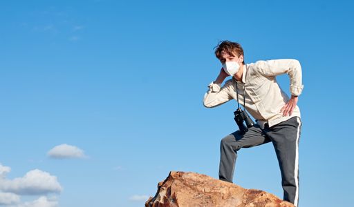 A side view of a Spanish young male wearing a facemask standing on a rock posing against the blue sky