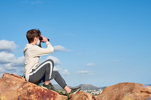 A shallow focus of an explorer from Spain in a white shirt sitting on a rock in the sight of a town looking through binoculars