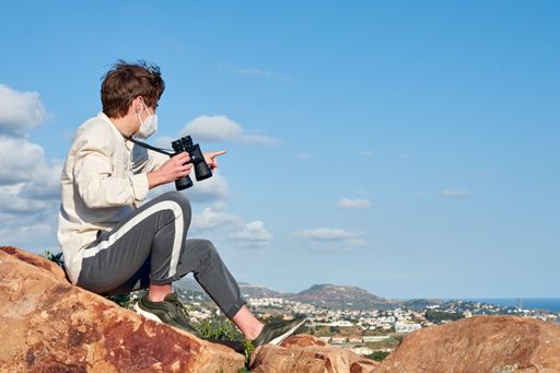 A shallow focus shot of a young explorer with binoculars from Spain pointing out mountains over a coastal town sitting on a rock