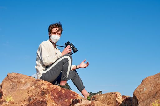 A Spanish adventurous male traveler wearing a mask sitting on a rock holding binoculars