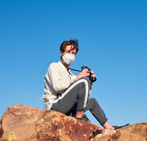 A front view of an explorer from Spain wearing a mask sitting on a rock looking into the camera on a sunny day