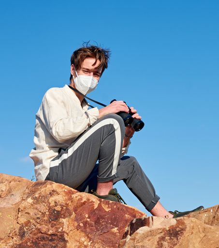 A front view of an explorer from Spain wearing a mask sitting on a rock smiling at the camera