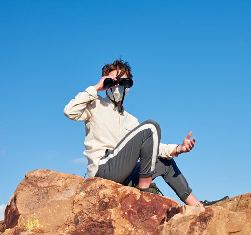 A front view of a young man from Spain wearing a white shirt and a face mask sitting on a rock, observing through binoculars-confused