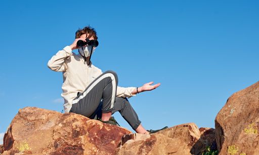 A shallow focus shot of a young man from Spain wearing a white shirt and a face mask sitting on a rock, observing through binoculars surprised