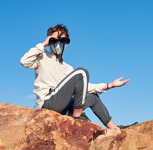 An adventurous young male traveler wearing a mask sitting on a rock and using binoculars