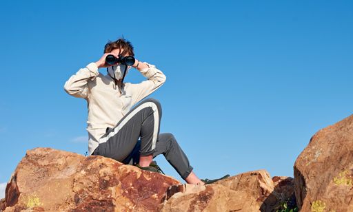 A shallow focus shot of a young man from Spain wearing a white shirt and a face mask sitting on a rock, observing through binoculars