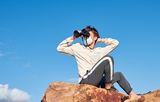 An adventurous young male traveler wearing a mask sitting on a rock and using binoculars