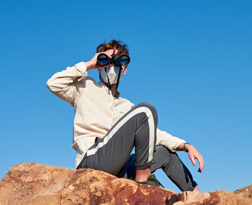 A front view of a young man from Spain wearing a white shirt and a face mask sitting on a rock, observing through binoculars