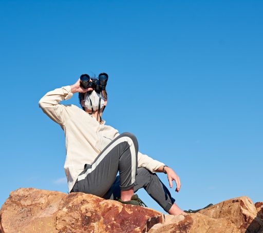 A shallow focus shot of a young man from Spain wearing a white shirt and a face mask sitting on a rock, observing through binoculars