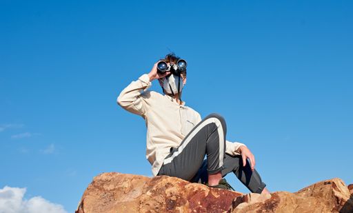 A front view of a young man from Spain wearing a white shirt and a face mask sitting on a rock, observing through binoculars