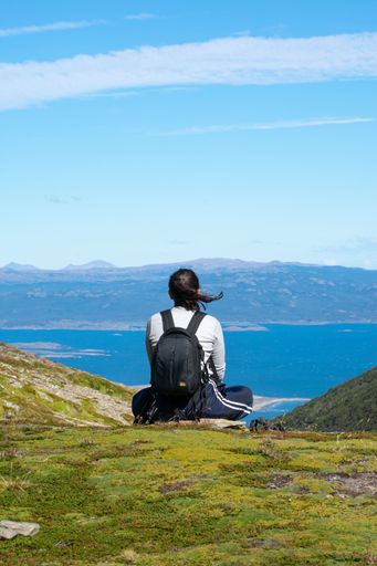 A young female trekker sitting on a lush green grassy area, taking in the stunning view of the vast blue ocean in front of her