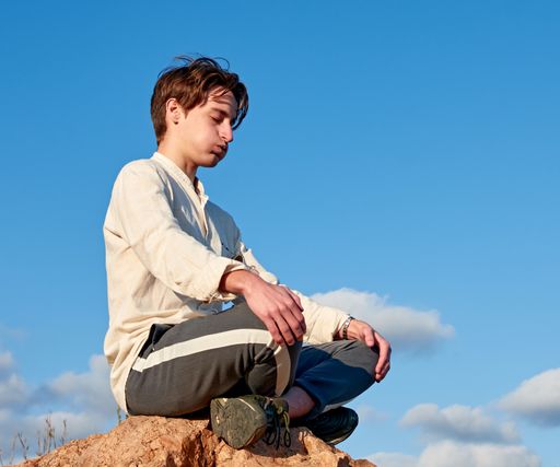 A shallow focus of a Spanish young male sitting on a rock and meditating against the cloudy blue sky
