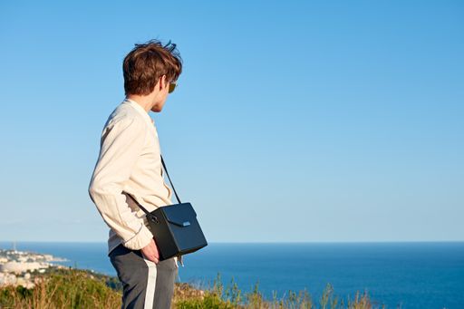 A Caucasian man from Spain standing on a grassy hill next to the ocean with a black purse over his shoulder