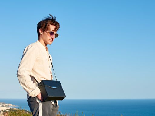 A Caucasian man from Spain standing on a grassy hill next to the ocean with a black purse over his shoulder
