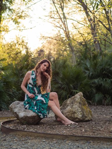 A vertical shot of a young female in a summer dress with leaves print sitting on a stone in a forest