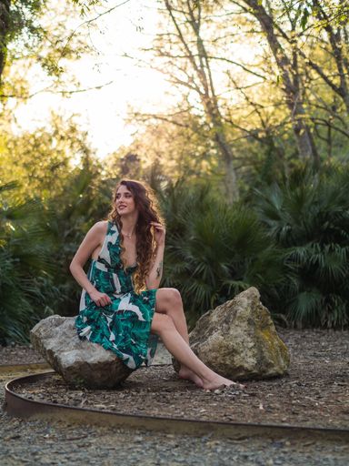 A vertical shot of a stylish female in a summer dress with leaves print sitting on a stone in a forest