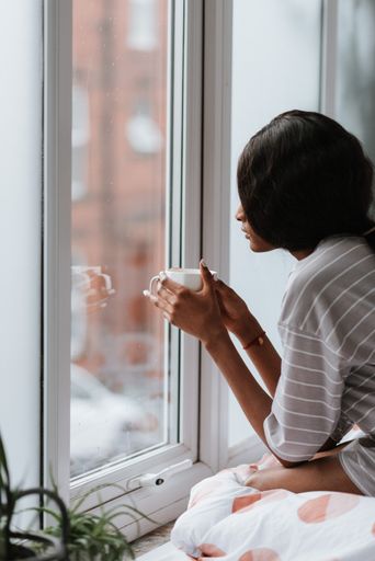A young African female with a cup of tea looking out of a window while sitting on the bed