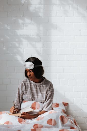 A thoughtful young African female with a sleeping eye mask on her head writing notes while sitting on the bed