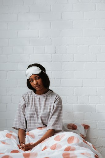 A thoughtful young African female with a white sleeping eye mask on her head sitting on the bed after waking up in the morning