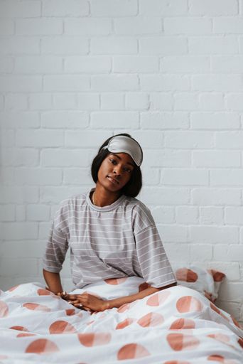 A thoughtful young African female with a white sleeping eye mask on her head sitting on the bed after waking up in the morning