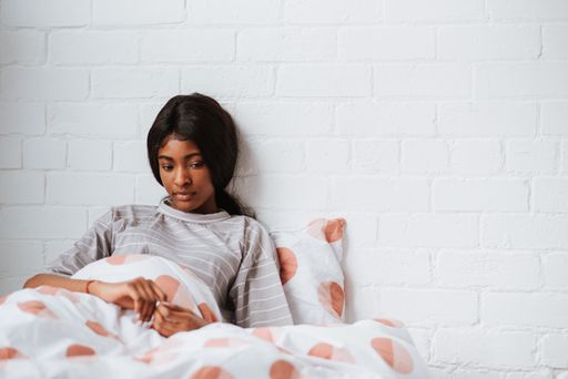 A sad young African female sitting on the bed leaning against a white wall after waking up in the morning