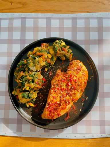A close-up image of a plated meal including a large grilled chicken breast served with a side dish of mixed vegetables