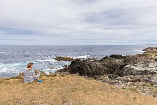 Caucasian blonde woman sitting on grass chatting on mobile phone by the sea. Woman in casual  listening music on cell phone on vacation. Copy space.