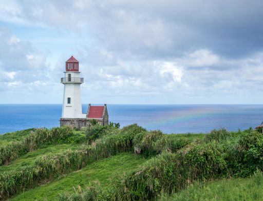 A majestic white lighthouse situated on top of a lush hill overlooking a rainbow and a calm sea