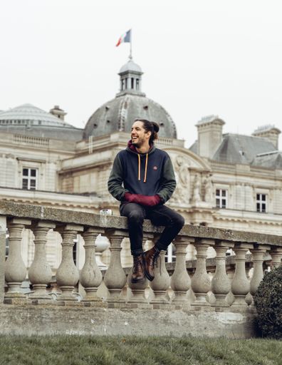 A Hispanic male sits on the edge of a stone fence with a typical Parisian building in the background