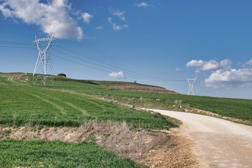 A landscape of dirt road between green fields with high voltage electrical pylons