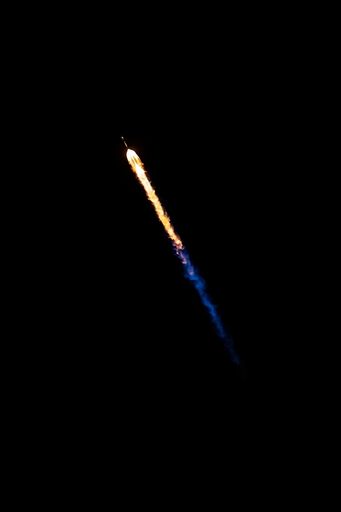 An illuminated space shuttle flying through the dark night sky, with a trail of smoke billowing behind it