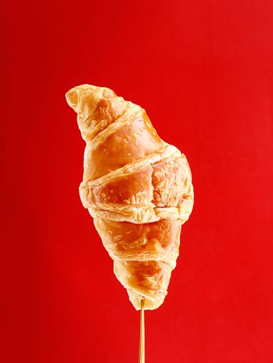 An isolated image of a golden-brown croissant skewered on a wooden stick against a bright red background