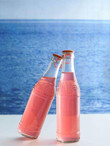 Two bottles of vibrant pink soda displayed on a table overlooking a beautiful beach