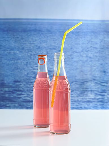 Two bottles of vibrant pink soda displayed on a table overlooking a beautiful beach