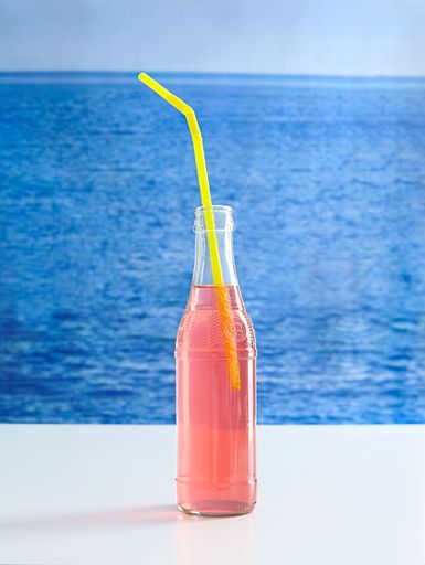A vibrant pink beverage in a clear bottle with a straw next to a tranquil ocean