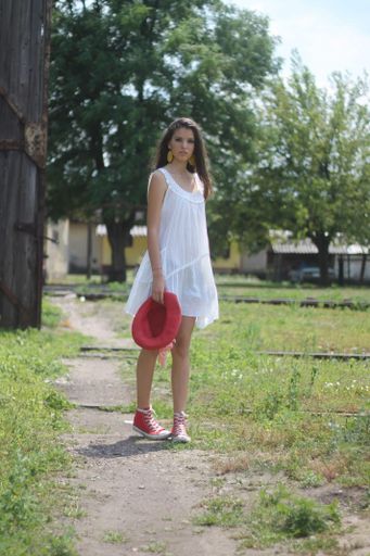 A vertical shot of a beautiful Caucasian brunette walking wearing a white dress holding a red hat