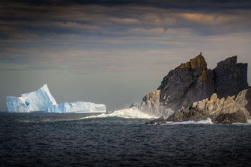A majestic iceberg seen in calm waters off of a rugged rocky cliff face