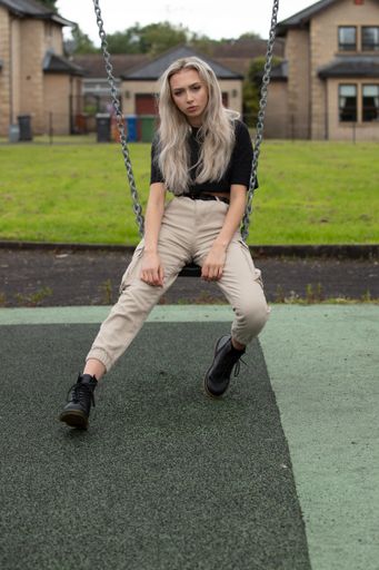 A vertical shot of a blonde girl sitting on the swing in the background of the buildings