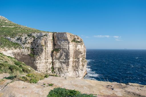 Limestone sheer cliffs showing layers of sedimentary rock next to coastline of Migra l-Ferha, Malta. Waves hitting the base of the cliffs and blue sky