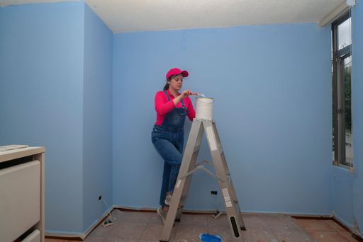 Beautiful woman dressed in overalls and red t-shirt going up a metal ladder with a jar of paint next to a blue wall in an empty room