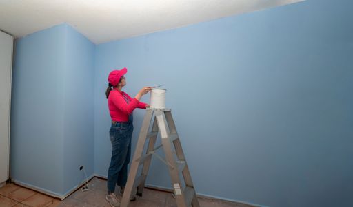 Beautiful woman dressed in overalls and red t-shirt going up a metal ladder with a jar of paint next to a blue wall in an empty room