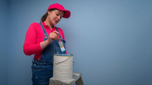 Beautiful woman dressed in overalls and red t-shirt standing on a metal ladder taking the brush out of the jar of blue paint next to a blue wall