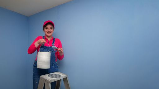 Beautiful woman dressed in overalls standing on a metal ladder, seen from the front, holding the brush and the paint jar next to a blue wall